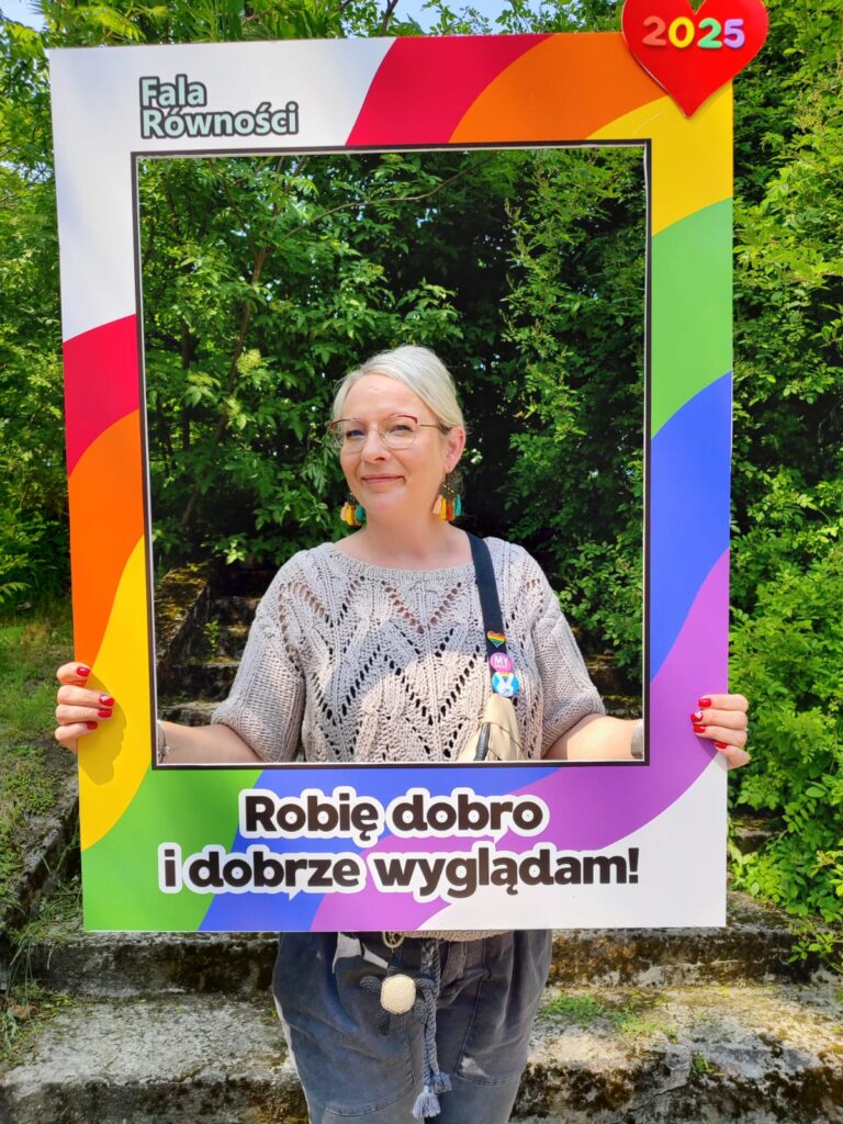 Uśmiechnięta kobieta trzyma w dłoniach tęczową ramkę otaczającą jej twarz z napisem Robię dobro i dobrze wyglądam! Ma okulary, kolorowe kolczyki, białe włosy i dzianinowy sweter.