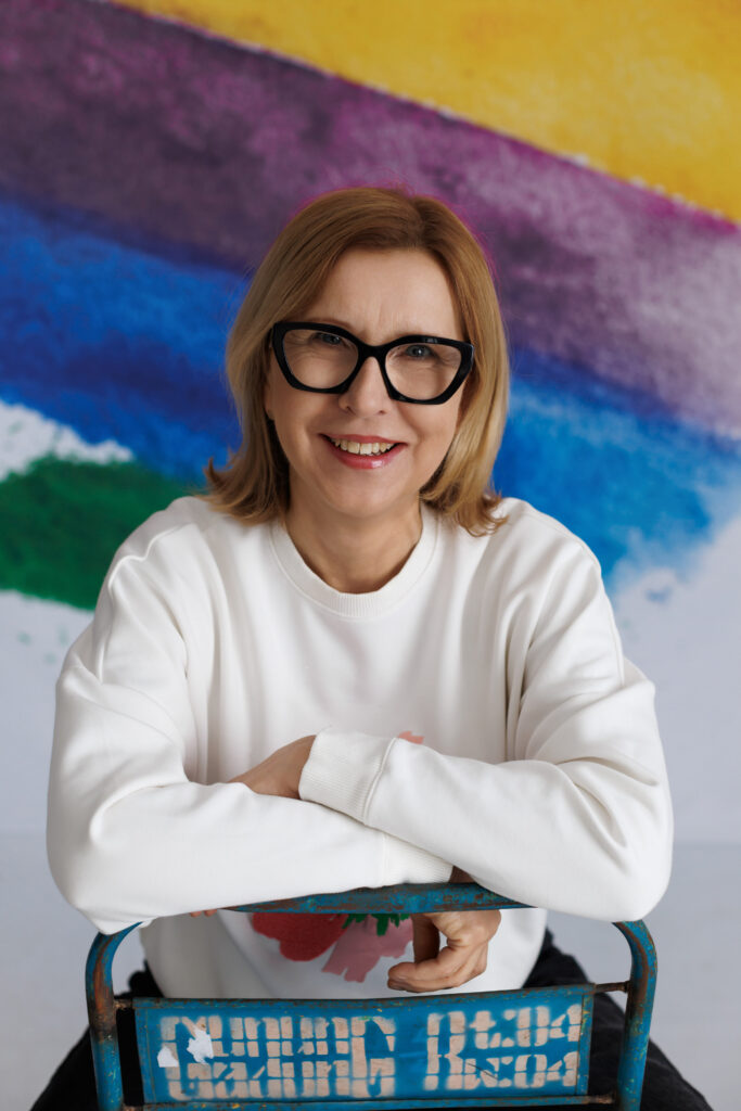 Blond osoba w okularach siedzi opierając się przodem rękoma na niebieskim krześle uśmiecha się szeroko do widza. W tle tęczowe serce.