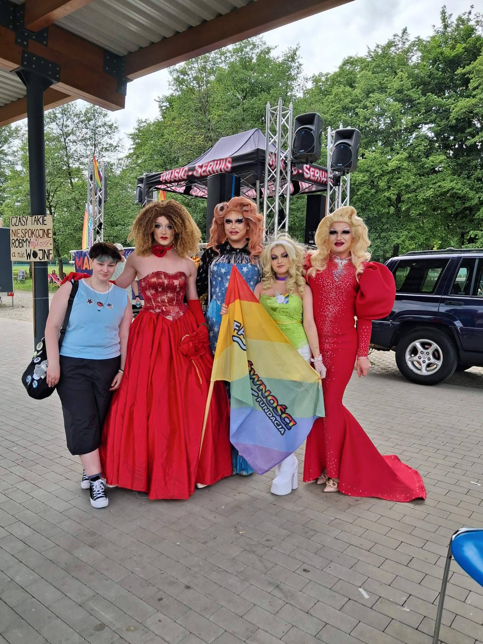 Zdjęcie grupowe wykonane na zewnątrz, na którym pozuje pięć osób: jedna osoba w stroju casualowym i cztery drag queens w efektownych kostiumach. Drag queens mają na sobie suknię w kolorze czerwonym, suknię w kolorze niebieskim ze wzorem flagi USA oraz limonkowe i fioletowe kreacje. Dwie z nich trzymają dużą, tęczową flagę z logo fundacji. Tło stanowią drzewa, nagłośnienie estradowe i zaparkowany samochód. Po lewej stronie widać transparent z napisem: "CZASY TAKIE NIESPOKOJNE RÓBMY miłość a nie WOJNĘ".