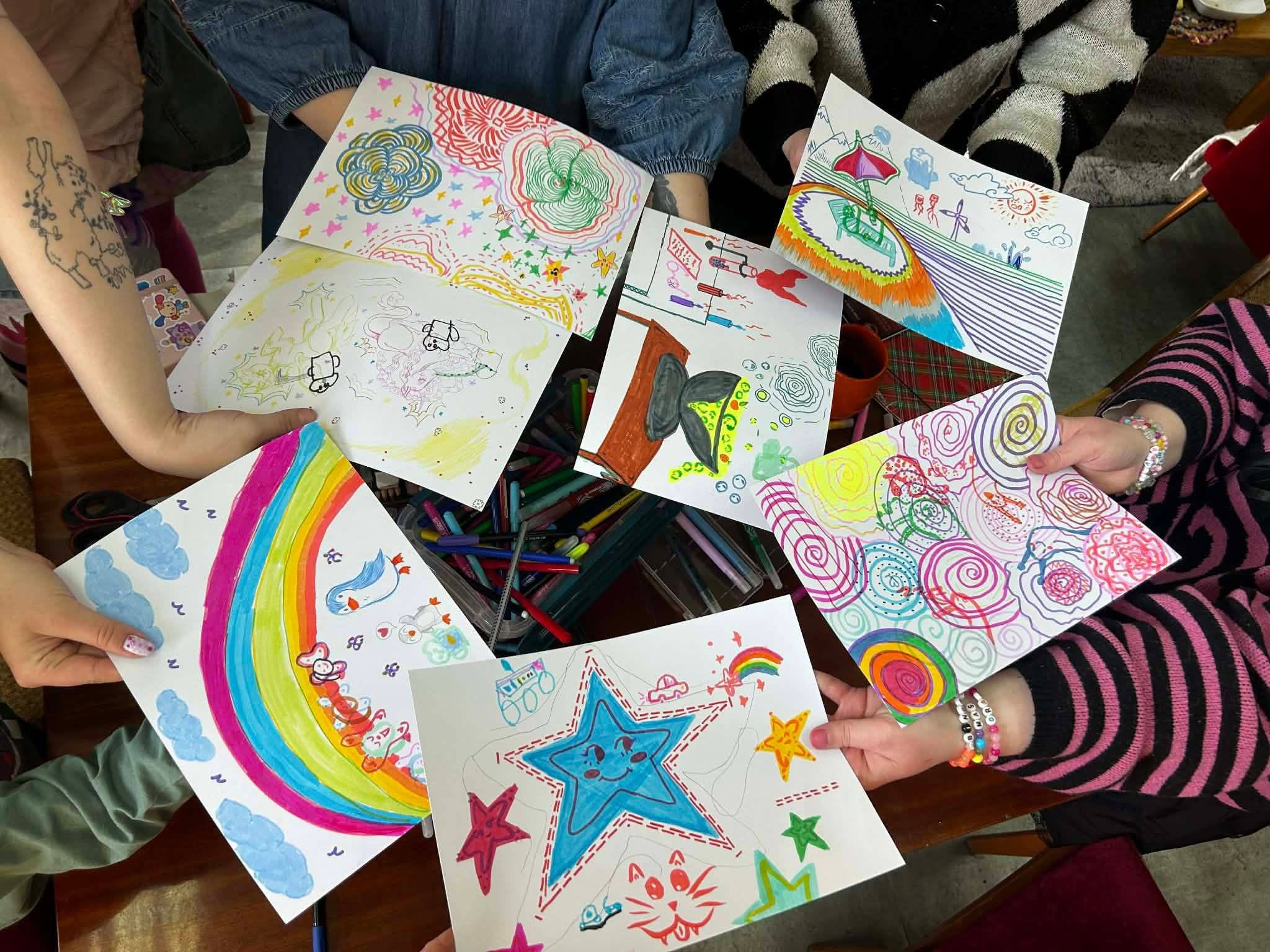 djęcie z perspektywy lotu ptaka (z góry) przedstawia warsztatowy, kreatywny moment. Kilka osób siedzi wokół drewnianego stołu, na którym w centralnej części leży duży pojemnik z mnóstwem kolorowych flamastrów. Dłonie uczestników tworzą okrąg, a każda z osób trzyma białą kartkę formatu A5 z własnoręcznie wykonanym rysunkiem. Wśród zaprezentowanych prac można dostrzec: Po lewej na dole: Rysunek z dużym motywem kolorowej tęczy, niebieskich chmur i małych ptaszków. Na dole w środku: Duża, uśmiechnięta niebieska gwiazda otoczona mniejszymi gwiazdkami, rysunkiem kota oraz samochodziku. Po prawej na dole: Abstrakcyjne, zakręcone spirale w kolorach różu, fioletu, żółci i zieleni. Po prawej na górze: Scena letnia z leżakiem pod parasolem plażowym, słońcem, chmurami i morzem w paski. Na górze w środku i po lewej: Prace z psychodelicznymi, hipnotyzującymi wzorami przypominającymi kwiaty oraz szkice drobnych postaci. Widoczne ręce należą do różnych osób – jedna z nich ma wytatuowane przedramię, inna ma na sobie pasiasty, czarno-różowy sweter, a na nadgarstkach niektórych uczestników lśnią kolorowe, koralikowe bransoletki. Kadr jest dynamiczny, radosny i pełen ekspresji.