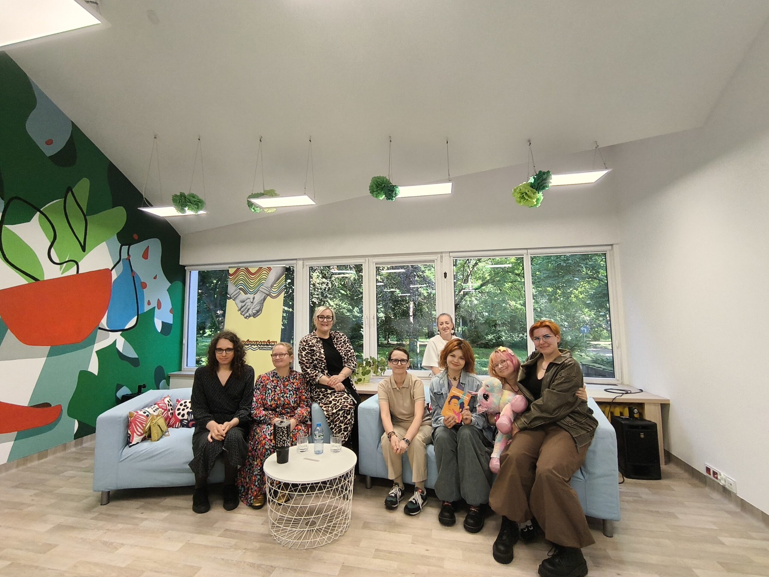 Zdjęcie przedstawia grupę ośmiu uśmiechniętych osób siedzących wspólnie w jasnym, nowocześnie urządzonym i przyjaznym wnętrzu. Pozujący siedzą blisko siebie na dwóch jasnoniebieskich sofach, a jedna z osób stoi za nimi. W tle widać duże okna wychodzące na zielony park oraz barwny, artystyczny mural na ścianie. Panuje swobodna, radosna i pełna ciepła atmosfera.