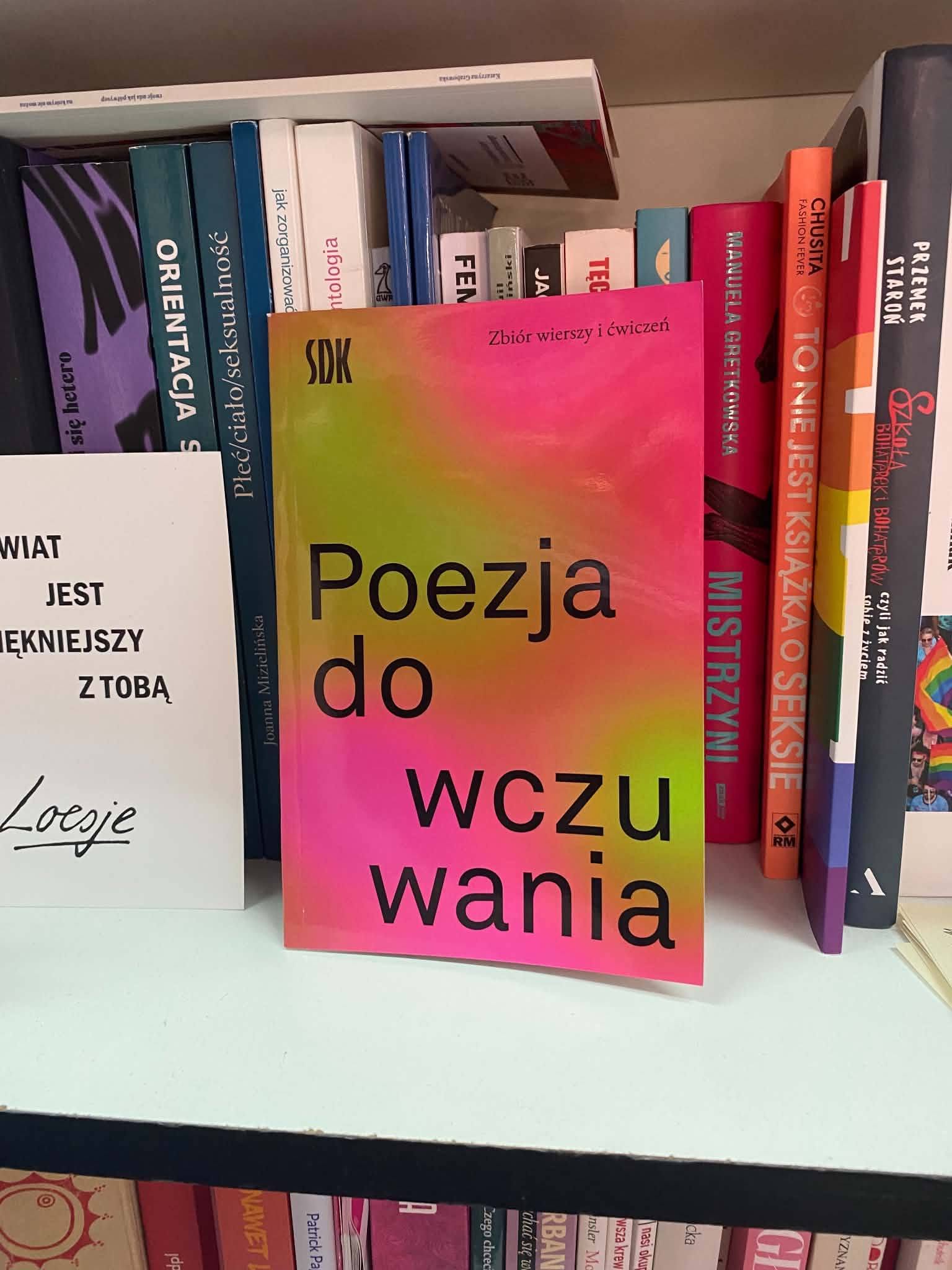 Zdjęcie przedstawia półkę z książkami, na której w centralnym miejscu wyeksponowano jedną książkę, opartą o inne woluminy. Na pierwszym planie znajduje się książka w miękkiej okładce o bardzo wyrazistej, neonywej kolorystyce – przejście tonalne od żółci, przez zieleń i fiolet, aż po intensywny róż. Na okładce widnieje duży, czarny napis: „Poezja do wczuwania”. Nad nim umieszczono małe logo „SDK” oraz mniejszy podtytuł: „Zbiór wierszy i ćwiczeń”. Po lewej stronie tej książki stoi biała karteczka lub tabliczka z czarnym napisem: „ŚWIAT JEST PIĘKNIEJSZY Z TOBĄ”, pod którym widnieje odręczny podpis „Locsje”. W tle za główną książką widoczne są grzbiety innych pozycji stojących na półce, m.in.: Przemek Staroń – Szkoła bohaterek i bohaterów Manuela Gretkowska – Mistrzyni Joanna Mizielińska – Płeć/ciało/seksualność Książki stoją na jasnej, białej półce regału, a poniżej widać fragment kolejnej półki wypełnionej grzbietami innych książek. Zdjęcie wykonano w jasnym, sztucznym lub dziennym świetle.
