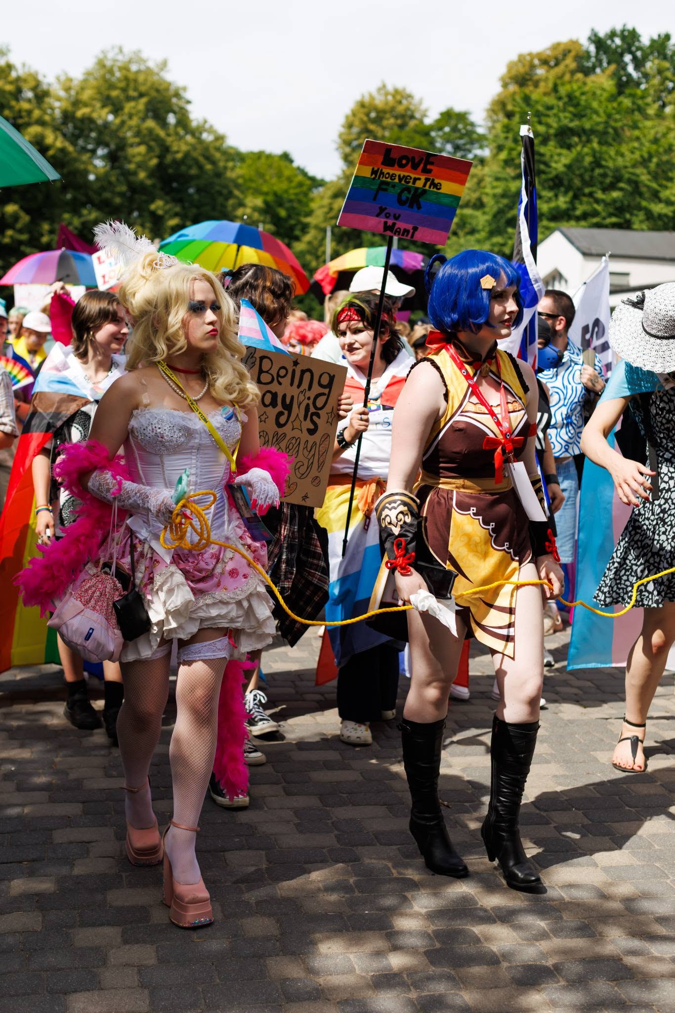Zdjęcie przedstawia tłum ludzi maszerujących ulicą. Jest to marsz równości osób LGBT+, widać liczne flagi i transparenty. Na pierwszym planie, w centrum uwagi, znajdują się dwie osoby w strojach cosplay. Z lewej strony idzie osoba przebrana za postać w stylu Lolita lub burleski. Ma puszystą, blond perukę spiętą w kok z piórem, mocny niebieski makijaż oczu i perłowy naszyjnik. Ubrana jest w biały, gorsetowy top zdobiony cekinami, białą mini spódniczkę z falbanami, białe kabaretki i jasnoróżowe buty na grubej platformie. W dłoniach trzyma jaskraworóżową, pierzastą boasę oraz gruby żółty sznur, a przez ramię ma przewieszoną różową torebkę. Z prawej strony, obok niej, maszeruje osoba w cosplayu Xiangling z gry "Genshin Impact". Ma charakterystyczną granatową perukę z upięciem, czerwoną czerwoną opaskę na szyi i brązowo-żółty strój stylizowany na chiński qipao z wycięciami. Nosi czarne kozaki do kolan. W dłoniach trzyma ten sam żółty sznur, co osoba obok. W tłumie za nimi widać wiele innych osób. Niektóre niosą transparenty. Jeden z nich, tekturowy, głosi: „Being gay is okay!” (Bycie gejem jest w porządku!). Inny, kolorowy, w barwach tęczy, ma napis: „LOVE whoever The FCK You Want” (Kochaj kogo kura chcesz). Ponad głowami maszerujących powiewają flagi, w tym tęczowa flaga dumy oraz biało-różowo-niebieska flaga osób transpłciowych. W tle widać zielone drzewa i fragmenty budynków pod jasnym niebem. Atmosfera wydaje się radosna i pełna energii.
