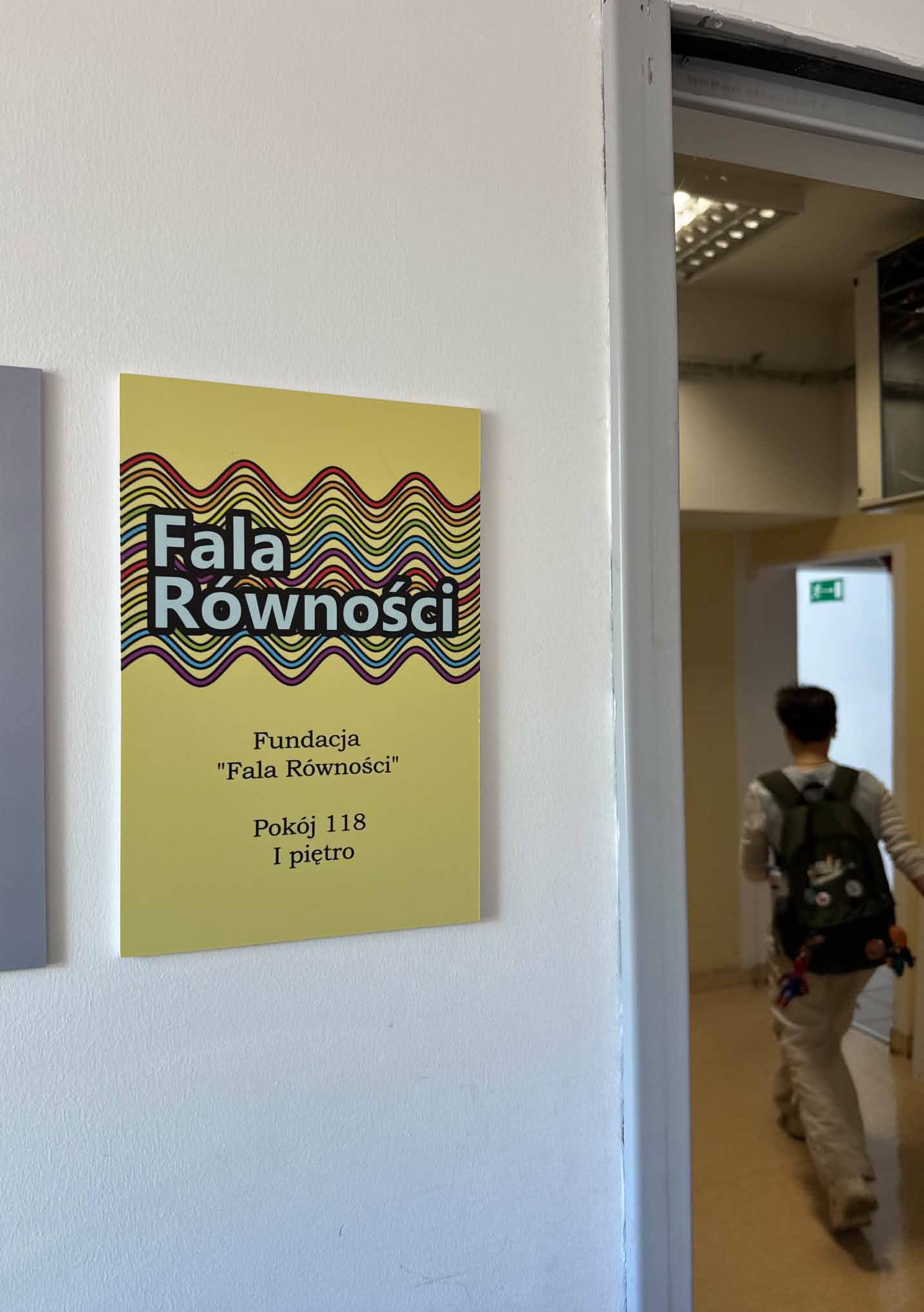 Zdjęcie przedstawia fragment białej, gładkiej ściany wewnątrz budynku, na której zamontowana jest tabliczka informacyjna. Po prawej stronie widać otwarty otwór drzwiowy z widokiem na korytarz. Głównym elementem zdjęcia jest prostokątna tabliczka w jasnym, żółtym kolorze. Jest ona zamocowana płasko na ścianie. W górnej części tabliczki znajduje się kolorowy wzór graficzny: nakładające się na siebie faliste linie w kolorach tęczy (m.in. czerwonym, żółtym, fioletowym i czarnym). Na tle tych fal, po lewej stronie, widnieje napis dużą, białą czcionką w dwóch rzędach: „Fala Równości”. Poniżej grafiki z falami, na żółtym tle tabliczki, znajduje się tekst wydrukowany ciemniejszą (prawdopodobnie czarną) czcionką. W pierwszym rzędzie napisane jest: „Fundacja”. W drugim rzędzie: „"Fala Równości"”. W trzecim rzędzie: „Pokój 118”. W czwartym, ostatnim rzędzie: „I piętro”. Na lewo od żółtej tabliczki widać fragment innej, gładkiej, jasnoszarej płyty lub tablicy bez widocznych napisów. W prawej części zdjęcia, przez otwarte drzwi bez framugi, widać jasny korytarz. W głębi korytarza oddala się osoba w ciemnej bluzie i jasnych spodniach, niosąca plecak (tyłem do obiektywu). Plecak ma kilka małych, jasnych, okrągłych elementów (być może przypinek lub odblasków) i kolorowe, zwisające paski. Na ścianie korytarza w tle widać mniejszą, zieloną tabliczkę, prawdopodobnie znak ewakuacyjny. Oświetlenie wewnątrz korytarza i na korytarzu w tle pochodzi z lamp sufitowych. Samo zdjęcie jest zrobione z perspektywy stojącej osoby, pod lekkim kątem do ściany, na której wisi tabliczka.