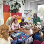 Ogólny zarys sceny Zdjęcie przedstawia kameralną, twórczą i przyjazną scenę wewnątrz jasnego, kolorowego pokoju (Tęczowej Miejscówki). Grupa sześciu osób zebrała się wokół dużego, drewnianego stołu konferencyjnego, na którym leżą przerabiane ubrania, materiały krawieckie, kłębki włóczki oraz biały pluszowy miś. Wszyscy wyglądają na zrelaksowanych i skupionych na wspólnym spędzaniu czasu. Pierwszy plan i stół: Na pierwszym planie (u dołu po lewej): Tyłem do obiektywu siedzi osoba z długimi, gęstymi, miedziano-rudymi włosami, ubrana w jasny, puszysty kardigan z frędzlami. Rozłożone ubranie na stole: Przed nią leżą rozpostarte niebieskie dżinsowe spodnie, które są w trakcie artystycznej przeróbki. Naszyto na nie różnorodne, kolorowe łaty i aplikacje: m.in. jasną koronkową łatę z kwiatem, czerwoną naszywkę z cekinami, różową kokardkę, szydełkowany czerwony trójkąt oraz białą aplikację z czaszką. Na spodniach leżą małe nożyczki krawieckie. Przybory na stole: Na gładkim, brązowym blacie stołu porozkładane są nożyczki, mulina, nici, poduszeczka na szpilki, przezroczysty worek wypełniony kolorowymi motkami wełny i ścinkami tkanin, kubek z herbatą, wstążka w tęczowych barwach oraz małe tekturowe pudełko. Osoby przy stole (od lewej do prawej): Osoba stojąca po lewej stronie: Pochyla się czule nad siedzącą obok koleżanką, opierając policzek o jej głowę. Ma bardzo kolorowe, pastelowe włosy (różowo-żółto-zielone) spięte spineczkami, jasną koszulkę w drobny wzór i fartuszek lub spódniczkę w kolorową szachownicę. Osoba siedząca po lewej (w centrum kadru): Uśmiecha się lekko w stronę aparatu. Ma krótkie, proste włosy w odcieniu intensywnego różu/bordo z prostą grzywką oraz okulary w różowych oprawkach. Ubrana jest w różowo-białą bluzkę w paski, na którą założony ma różowy top, a jej nadgarstki i szyję zdobi mnóstwo kolorowych koralików i naszyjników. Trzyma w dłoniach telefon lub drobny przedmiot. Osoba siedząca pośrodku: Młoda osoba z krótkimi, ciemnymi włosami, w ciemnoczerwonej koszulce polo w paski. Jest skupiona, z pochyloną głową przegląda lub rozwija materiały z foliowego worka. Kobieta stojąca po prawej stronie stołu (prowadząca, Weronika Teplicka): Stoi wyprostowana, uśmiecha się ciepło do aparatu. Ma ciemne włosy do ramion, okulary w ciemnych oprawkach i ubrana jest w soczyście zielony, rozpinany kardigan narzucony na czarną bluzkę. Biały pluszowy miś: Siedzi na stole tuż przed kobietą w zielonym swetrze. To nieduży, kremowo-biały miś z przewiązaną pod szyją niebieską wstążką. Osoba siedząca po prawej stronie: Siedzi bokiem do stołu na krześle z bordowym obiciem. Ma długie, ciemne, mocno kręcone włosy, ubrana jest w niebieską spódnicę w zielone kropki oraz ciemny, melanżowy płaszcz lub gruby sweter. Tło i przestrzeń: Po lewej stronie: Wysoki biały regał gęsto zastawiony książkami, komiksami (m.in. widać tomy mangi i powieści młodzieżowe) oraz grami planszowymi. Obok stoi wysoka szafa z frontami w jaskrawych, tęczowych kolorach (czerwonym i pomarańczowym), na której stoi donica z dużą, zieloną monsterą o liściach pnących się ku górze. W głębi: Za stołem stoi tablica typu flipchart / tablica magnetyczna, gęsto oklejona dziecięcymi i młodzieżowymi rysunkami, plakatami (widać m.in. jednorożce, napisy „LOVE IS LOVE!”, tęcze i zasady panujące w przestrzeni). Po prawej stronie: Duże okno z białymi żaluzjami, wychodzące na korony drzew. Obok okna fragment ściany lub filara ozdobiony pionową mozaiką z drobnych, kolorowych płytek. Całe wnętrze sprawia wrażenie bezpiecznego, ciepłego, domowego i pełnego twórczego zapału.