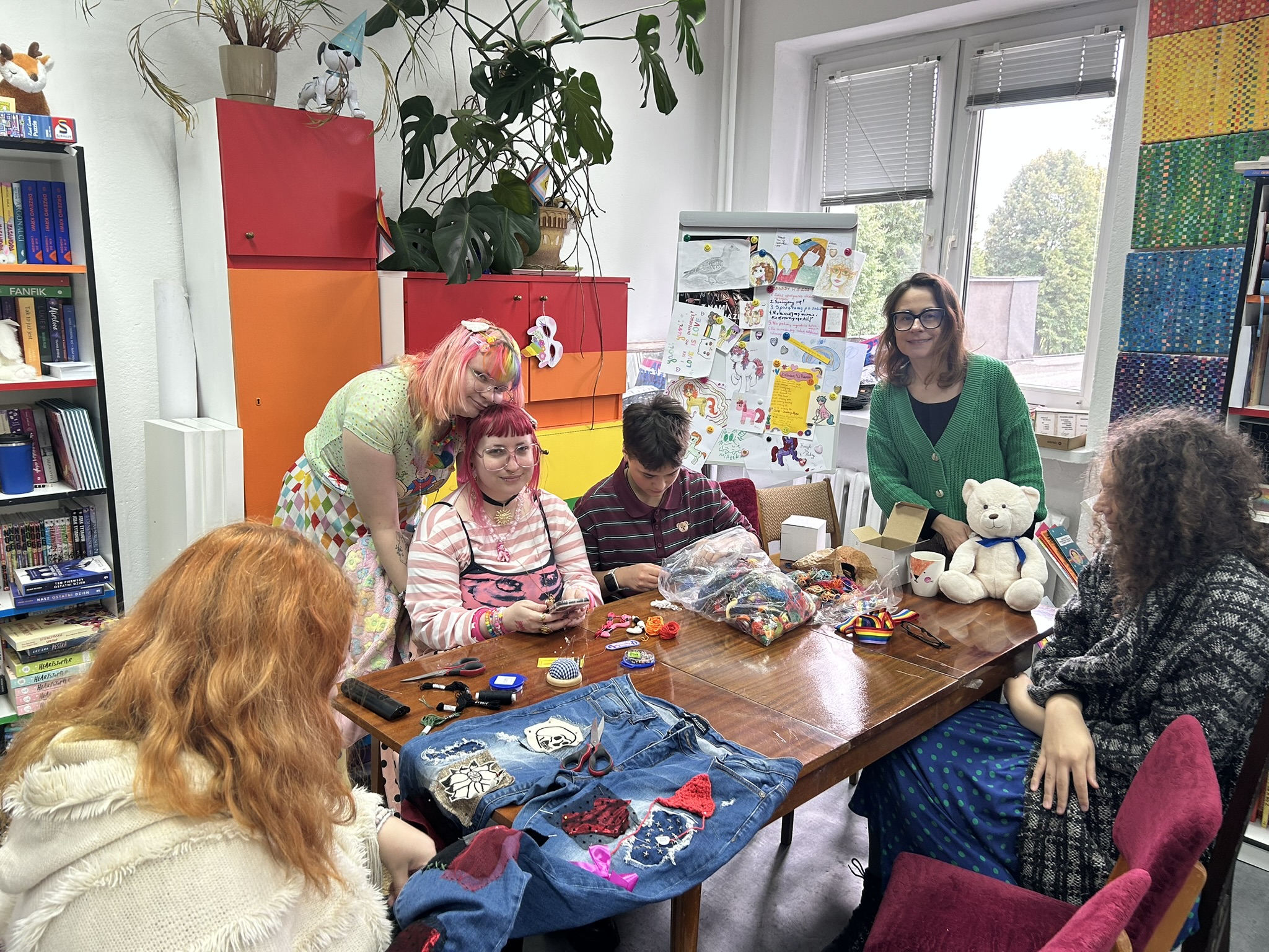 Ogólny zarys sceny Zdjęcie przedstawia kameralną, twórczą i przyjazną scenę wewnątrz jasnego, kolorowego pokoju (Tęczowej Miejscówki). Grupa sześciu osób zebrała się wokół dużego, drewnianego stołu konferencyjnego, na którym leżą przerabiane ubrania, materiały krawieckie, kłębki włóczki oraz biały pluszowy miś. Wszyscy wyglądają na zrelaksowanych i skupionych na wspólnym spędzaniu czasu. Pierwszy plan i stół: Na pierwszym planie (u dołu po lewej): Tyłem do obiektywu siedzi osoba z długimi, gęstymi, miedziano-rudymi włosami, ubrana w jasny, puszysty kardigan z frędzlami. Rozłożone ubranie na stole: Przed nią leżą rozpostarte niebieskie dżinsowe spodnie, które są w trakcie artystycznej przeróbki. Naszyto na nie różnorodne, kolorowe łaty i aplikacje: m.in. jasną koronkową łatę z kwiatem, czerwoną naszywkę z cekinami, różową kokardkę, szydełkowany czerwony trójkąt oraz białą aplikację z czaszką. Na spodniach leżą małe nożyczki krawieckie. Przybory na stole: Na gładkim, brązowym blacie stołu porozkładane są nożyczki, mulina, nici, poduszeczka na szpilki, przezroczysty worek wypełniony kolorowymi motkami wełny i ścinkami tkanin, kubek z herbatą, wstążka w tęczowych barwach oraz małe tekturowe pudełko. Osoby przy stole (od lewej do prawej): Osoba stojąca po lewej stronie: Pochyla się czule nad siedzącą obok koleżanką, opierając policzek o jej głowę. Ma bardzo kolorowe, pastelowe włosy (różowo-żółto-zielone) spięte spineczkami, jasną koszulkę w drobny wzór i fartuszek lub spódniczkę w kolorową szachownicę. Osoba siedząca po lewej (w centrum kadru): Uśmiecha się lekko w stronę aparatu. Ma krótkie, proste włosy w odcieniu intensywnego różu/bordo z prostą grzywką oraz okulary w różowych oprawkach. Ubrana jest w różowo-białą bluzkę w paski, na którą założony ma różowy top, a jej nadgarstki i szyję zdobi mnóstwo kolorowych koralików i naszyjników. Trzyma w dłoniach telefon lub drobny przedmiot. Osoba siedząca pośrodku: Młoda osoba z krótkimi, ciemnymi włosami, w ciemnoczerwonej koszulce polo w paski. Jest skupiona, z pochyloną głową przegląda lub rozwija materiały z foliowego worka. Kobieta stojąca po prawej stronie stołu (prowadząca, Weronika Teplicka): Stoi wyprostowana, uśmiecha się ciepło do aparatu. Ma ciemne włosy do ramion, okulary w ciemnych oprawkach i ubrana jest w soczyście zielony, rozpinany kardigan narzucony na czarną bluzkę. Biały pluszowy miś: Siedzi na stole tuż przed kobietą w zielonym swetrze. To nieduży, kremowo-biały miś z przewiązaną pod szyją niebieską wstążką. Osoba siedząca po prawej stronie: Siedzi bokiem do stołu na krześle z bordowym obiciem. Ma długie, ciemne, mocno kręcone włosy, ubrana jest w niebieską spódnicę w zielone kropki oraz ciemny, melanżowy płaszcz lub gruby sweter. Tło i przestrzeń: Po lewej stronie: Wysoki biały regał gęsto zastawiony książkami, komiksami (m.in. widać tomy mangi i powieści młodzieżowe) oraz grami planszowymi. Obok stoi wysoka szafa z frontami w jaskrawych, tęczowych kolorach (czerwonym i pomarańczowym), na której stoi donica z dużą, zieloną monsterą o liściach pnących się ku górze. W głębi: Za stołem stoi tablica typu flipchart / tablica magnetyczna, gęsto oklejona dziecięcymi i młodzieżowymi rysunkami, plakatami (widać m.in. jednorożce, napisy „LOVE IS LOVE!”, tęcze i zasady panujące w przestrzeni). Po prawej stronie: Duże okno z białymi żaluzjami, wychodzące na korony drzew. Obok okna fragment ściany lub filara ozdobiony pionową mozaiką z drobnych, kolorowych płytek. Całe wnętrze sprawia wrażenie bezpiecznego, ciepłego, domowego i pełnego twórczego zapału.