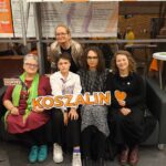 Na fotografii znajduje się pięć osób związanych z Falą Równości, które wspólnie, w radosnej atmosferze, pozują do zdjęcia na stoisku w przeszklonym holu Filharmonii Koszalińskiej. Cztery osoby siedzą ciasno obok siebie na ciemnej, skórzanej kanapie, a jedna stoi tuż za nimi, opierając się o oparcie. Od lewej siedzą: Starsza kobieta w okularach o ciemnych oprawkach i krótkich, szpakowatych włosach. Ma na sobie bordową tunikę, wzorzyste getry i turkusowe buty sportowe. Szyję ma owiniętą długim, jasnozielonym szalem, na którym widać kolorowe, masywne korale. W prawej dłoni trzyma małą tabliczkę na patyku z napisem „Kocham działać!” w pomarańczowo-białych barwach. Lewą ręką podtrzymuje duży napis „KOSZALIN”. Młoda osoba o bardzo krótkich, ciemnych włosach, w okularach, ubrana w białą koszulę z krawatem o fioletowym wzorze, ciemne spodnie i jasne sportowe buty. Siedzi pochylona lekko do przodu, a w rękach trzyma początek dużej, poziomej tablicy z napisem „KOSZALIN”. Osoba z długimi, ciemnymi, kręconymi włosami i w okularach, ubrana w długą, wzorzystą, czarno-szarą sukienkę, ciemne rajstopy i czarne buty sportowe. Siedzi pośrodku, trzymając drugą część tablicy z napisem „KOSZALIN”. Kobieta po prawej stronie o długich, lekko kręconych, jasnobrązowych włosach, naturalnie i ciepło uśmiechająca się do obiektywu. Jest ubrana w czarną bluzkę, czarną spódnicę i ciemny kardigan, a na nogach ma sznurowane, bordowe, skórzane buty (glany). W lewej dłoni, na wysokości klatki piersiowej, trzyma małe, pomarańczowe papierowe serduszko. Za siedzącymi, pośrodku, stoi: 5. Kobieta w okularach o spiętych, jasnych włosach, ubrana w błyszczącą, cekinową lub metaliczną narzutkę w odcieniu starego złota i ciemny t-shirt. Opiera się o kanapę i patrzy w obiektyw z łagodnym uśmiechem. Centralnym punktem zdjęcia jest trzymany przez siedzących duży, biały szyld z wyciętym, pomarańczowym napisem „KOSZALIN” w fioletowej obwódce. Tło: Za grupą znajduje się przeszklona ściana oraz metalowa konstrukcja stoiska, na której u góry i w tle wiszą duże, jasne plakaty zapowiadające koncerty w Filharmonii Koszalińskiej (z nazwiskami artystów i datami). Przez szyby w tle widać wnętrze budynku, kręcących się ludzi (w tym osobę w żółtej kurtce) oraz fragmenty wyposażenia, takie jak okrągły stolik barowy po prawej stronie. Na podłodze widać ciemne, wielkoformatowe kafelki. Zdjęcie jest naturalne, pełne ciepłych barw i bije z niego pozytywna, zespołowa energia